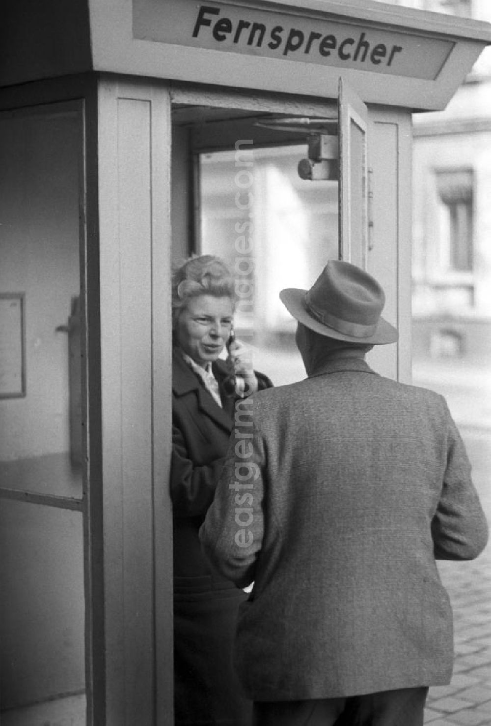 GDR photo archive: Leipzig - Eine Frau telefoniert in einem Telefonhäuschen in Leipzig mit einem öffentlichen Fernsprecher, ihr Mann hört an der geöffneten Tür mit zu.