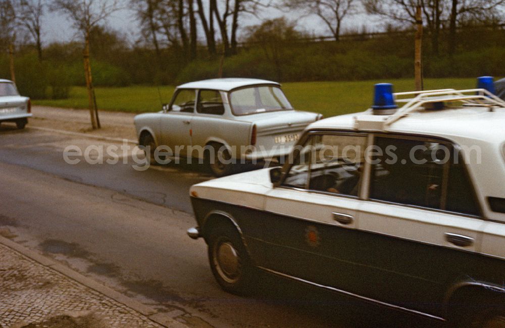 GDR image archive: Berlin - People's Police car drives on a street in East Berlin in the territory of the former GDR, German Democratic Republic. Trabant cars are parked on the side of the road