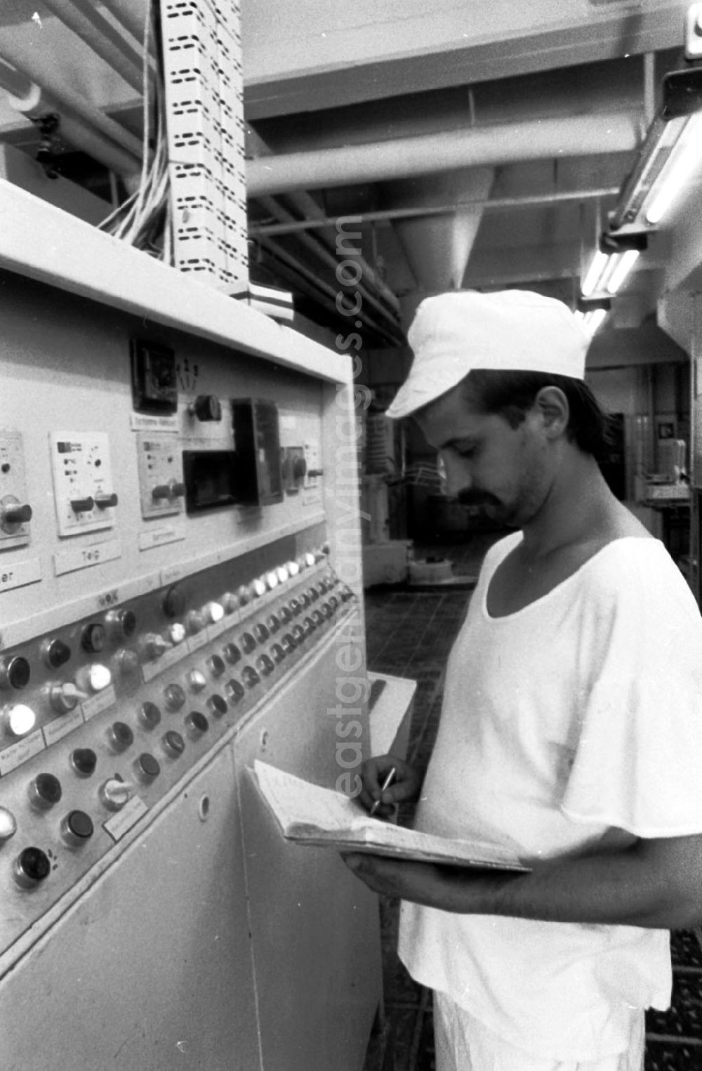 GDR photo archive: Berlin - Ein Techniker in einer Bäckerei in Heinersdorf steht vor Fließbandtechnik / Machine und schreibt Daten in Notizbuch.