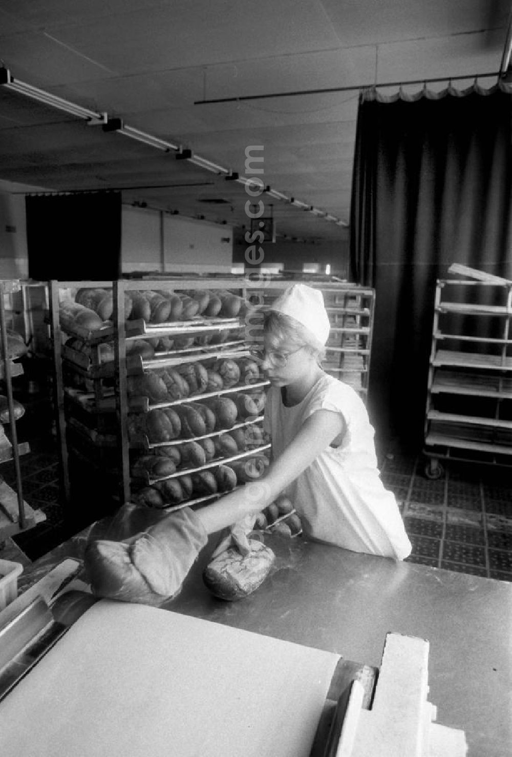 GDR picture archive: Berlin - Eine Arbeiterin einer Bäckerei in Berlin-Heinersdorf nimmt die fertig gebackenen und noch heißen Brote / Brotlaibe vom Fließband. Im Hintergrund sind Mischbrote im Regal gestapelt.