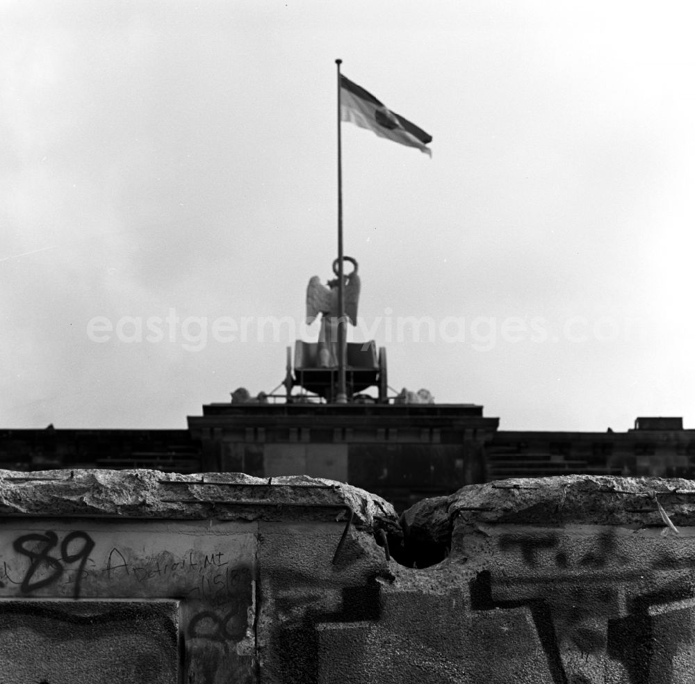 GDR picture archive: Berlin - View of the Quadriga on the Brandenburg Gate. In the wind Flag of the German Democratic Republic blows with a hammer, compasses and wreath of grain ears in Berlin. In the foreground is a hole in the wall crest of the Berlin Wall