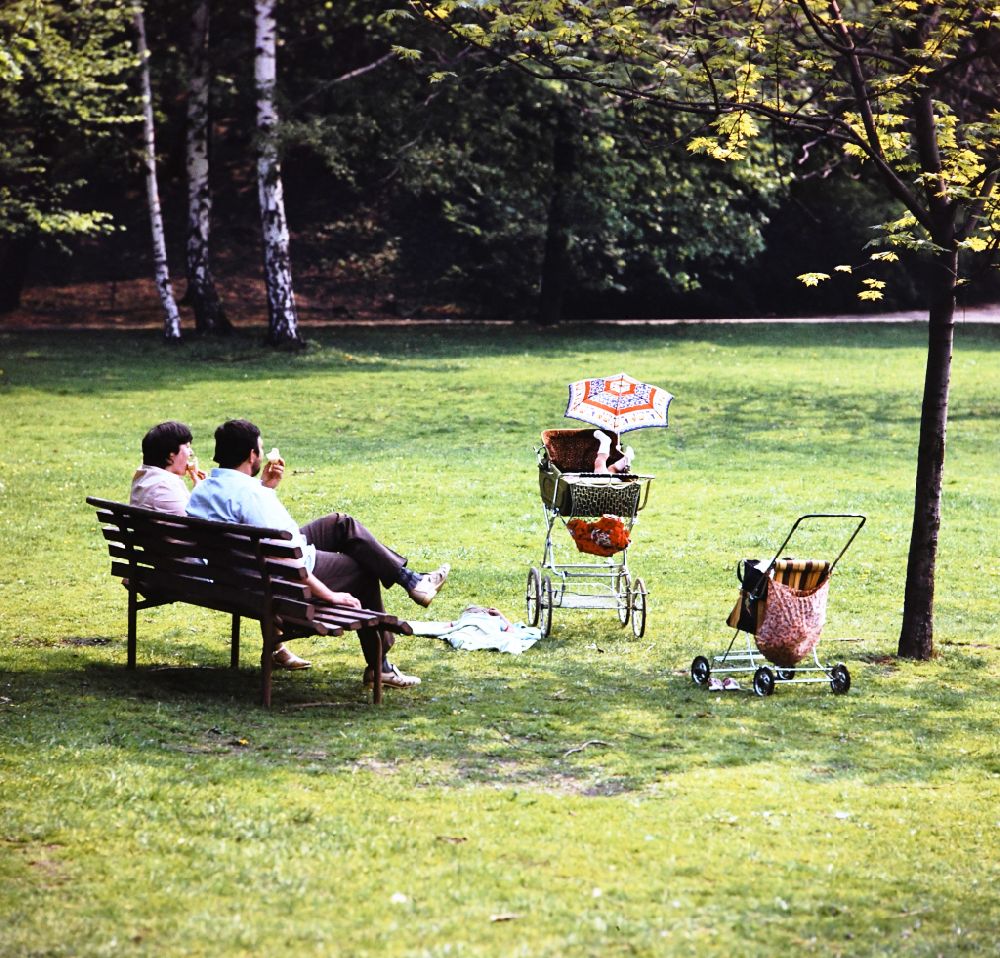 GDR image archive: Berlin - Family with pram or buggy sitting together on a park bench eating an ice cream in the Volkspark Friedrichshain in Eastberlin on the territory of the former GDR, German Democratic Republic