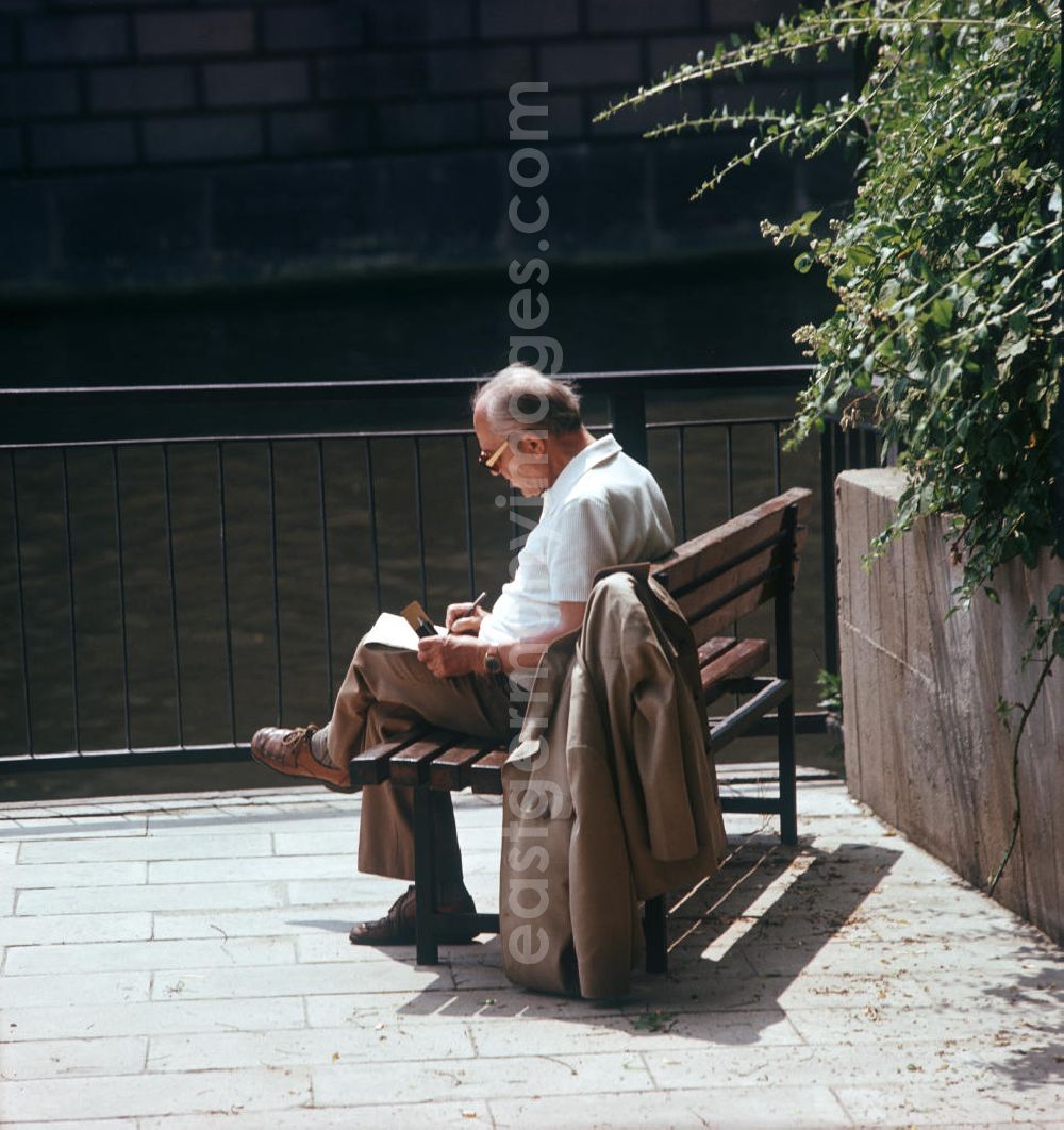 GDR picture archive: Berlin - Ein Mann sitzt auf einer Parkbank am Spreeufer und löst in einer Zeitung Kreuzworträtsel.