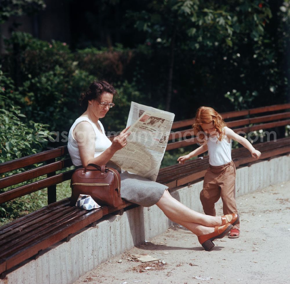 GDR image archive: Berlin - Eine Frau liest auf einer Parkbank ihre Zeitung, während ihre Tochter daneben spielt. Auf der Bank liegt ein Tetra Pak mit Trink-Vollmilch.