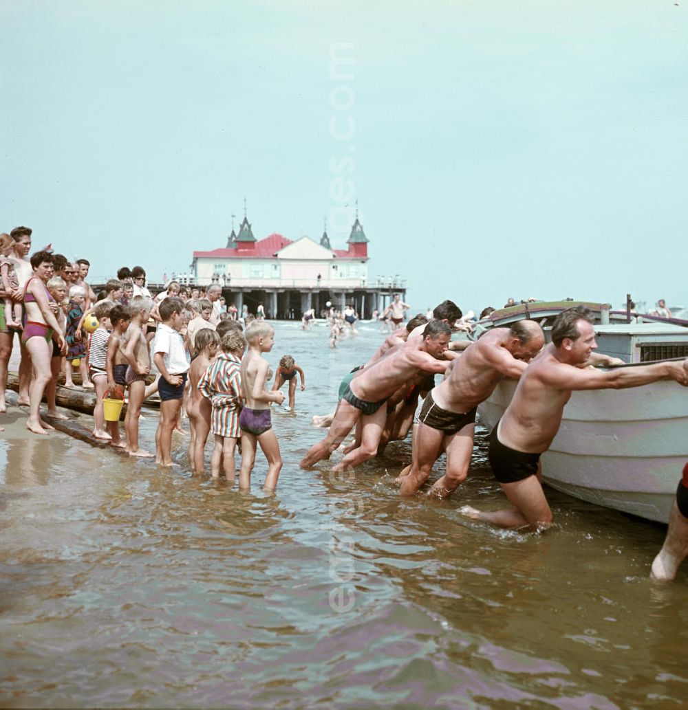 GDR image archive: Ahlbeck - Unter den neugierigen Augen anderer Badegäste schieben Männer ein Boot ins Wasser der Ostsee, im Hintergrund die Seebrücke Ahlbeck auf der Insel Usedom.