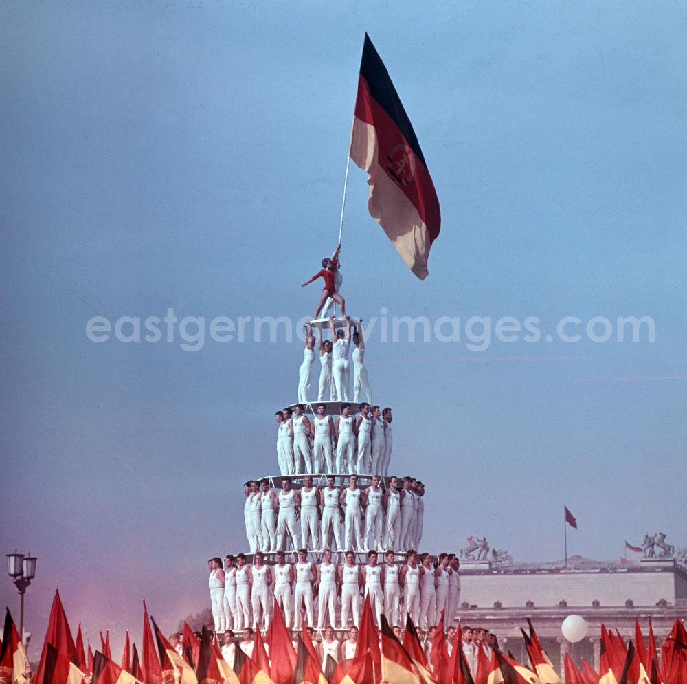 GDR picture archive: Leipzig - Sportler bauen eine Pyramide aus Menschen auf der Festwiese des Leipziger Zentralstadions beim V. Turn- und Sportfestes der DDR vom 24. bis 27.7.1969. Das Turnerpaar auf der Spitze der Pyramide hält die Fahne der DDR in die Höhe.