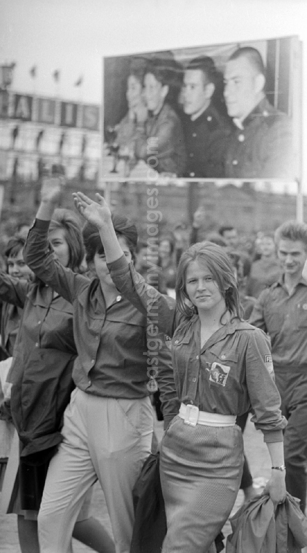 GDR image archive: Berlin - Zu Pfingsten 1964, vom 16.-18. Mai, findet in Berlin das letzte Deutschlandtreffen der Jugend für Frieden und Völkerfreundschaft statt. Hier Teilnehmer einer Demonstration. Eine junge Frau / Mädchen mit FDJ-Hemd und zwei andere Mitstreiterinnen hebt den Arm zum Gruß. Im Hintergrund eine Bild-tafel / Transparent mit dem Gesichtern sozialistischer Prominenter. Ziel der ersten Veranstaltung im Mai 1950 war, die kommunistische Jugend in Ost und West mobil zu machen und der damals in der BRD noch zugelassenen FDJ neue Anh?nger zuzuführen. 1951 wurde das Verbot gegen die FDJ in der BRD ausgesprochen. Nach 1964 entsprach eine gesamtdeutsche kommunistische Politik nicht mehr den Zielen der DDR-Regierung. In den 70er und 8