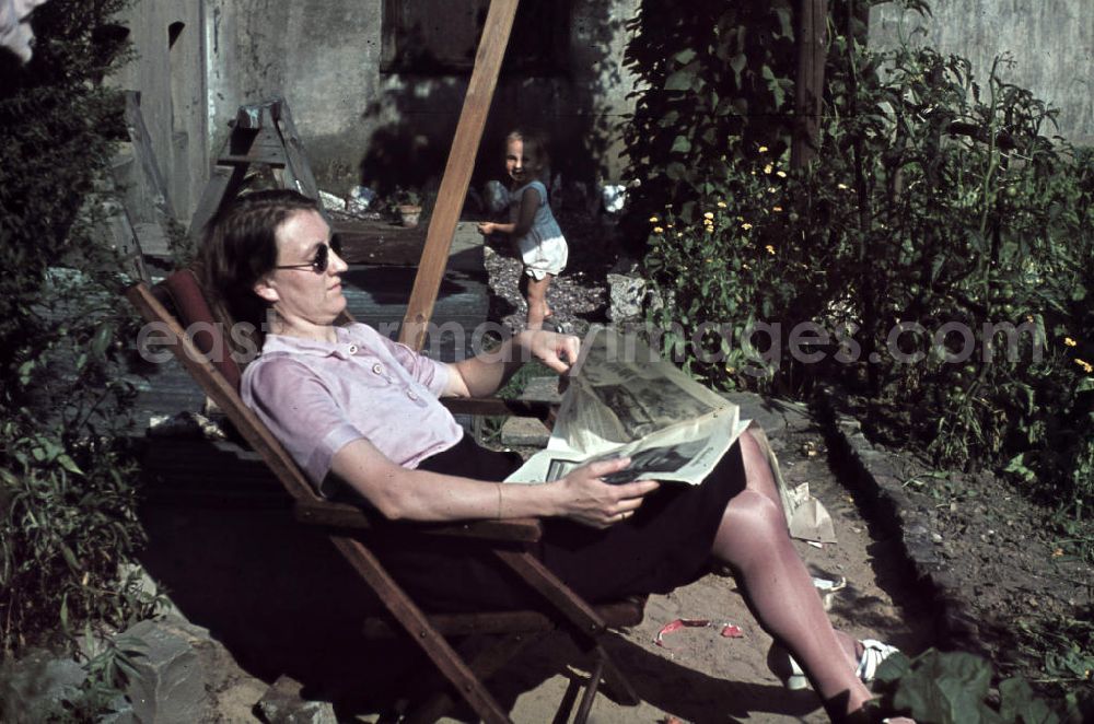 GDR picture archive: Siegen - Kleinkind spielt im Garten, die Mutter sitzt im Liegestuhl und liest eine Zeitung. Plying infant in a garden. The mother is sitting in a deck chair and read a newspaper.