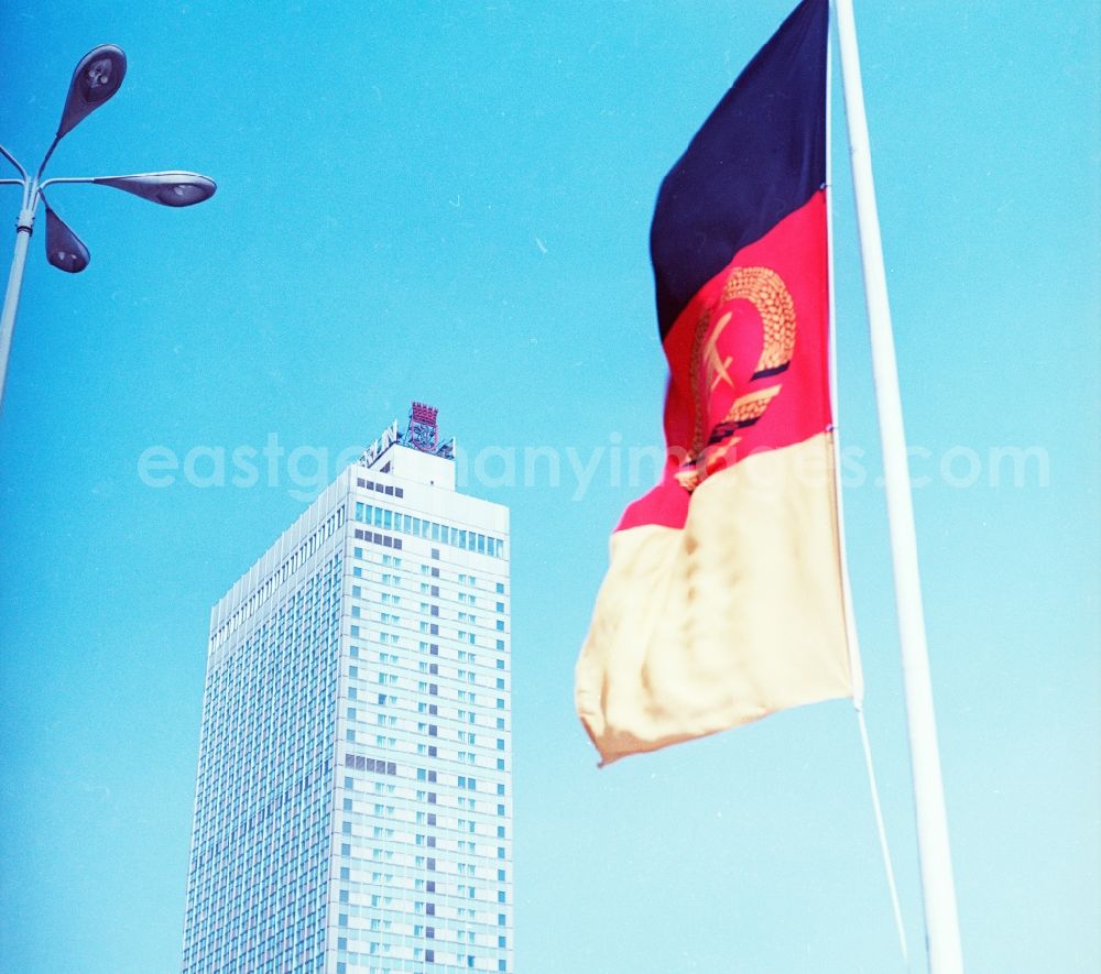 GDR photo archive: Berlin - Flag of the German Democratic Republic in Berlin. It shows the colors of the Weimar Republic. On the colors black, red and gold is in the middle the coat of arms of the GDR with hammer, compasses and wreath of grain ears. In the background is the Hotel Stadt Berlin and the Berlin coat of arms, the Berlin Bear