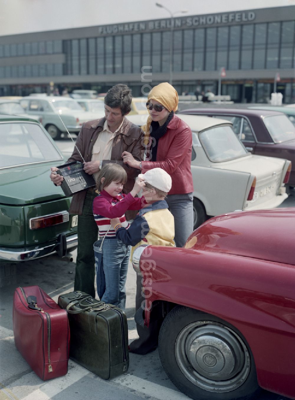 Schönefeld: Family between cars like Wartburg and Trabant in the parking lot in front of the airport Schoenefeld in the state Brandenburg on the territory of the former GDR, German Democratic Republic. Advertising shot for the radio cassette recorder Anett IS of the VEB Antennenwerke Bad Blankenburg
