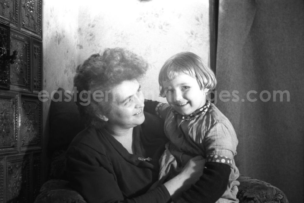 GDR picture archive: Leipzig - Frau mit einem Mädchen, welches auf ihrem Schoß sitzt. Innenaufnahme mit Polstersessel und Ausschnitt von einem Kachelofen. Bestmögliche Qualität nach Vorlage!