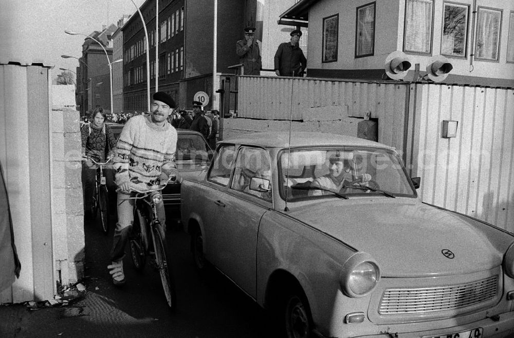 GDR photo archive: Berlin - Mauerfall / Grenzübergang Invalidenstraße, Bürger fahren mit Fahrrädern und Autos vom Typ Trabant durch Tor am Grenzübergang. Polizisten / Volkspolizisten stehen auf Erhöhung im Hintergrund und schauen zu.