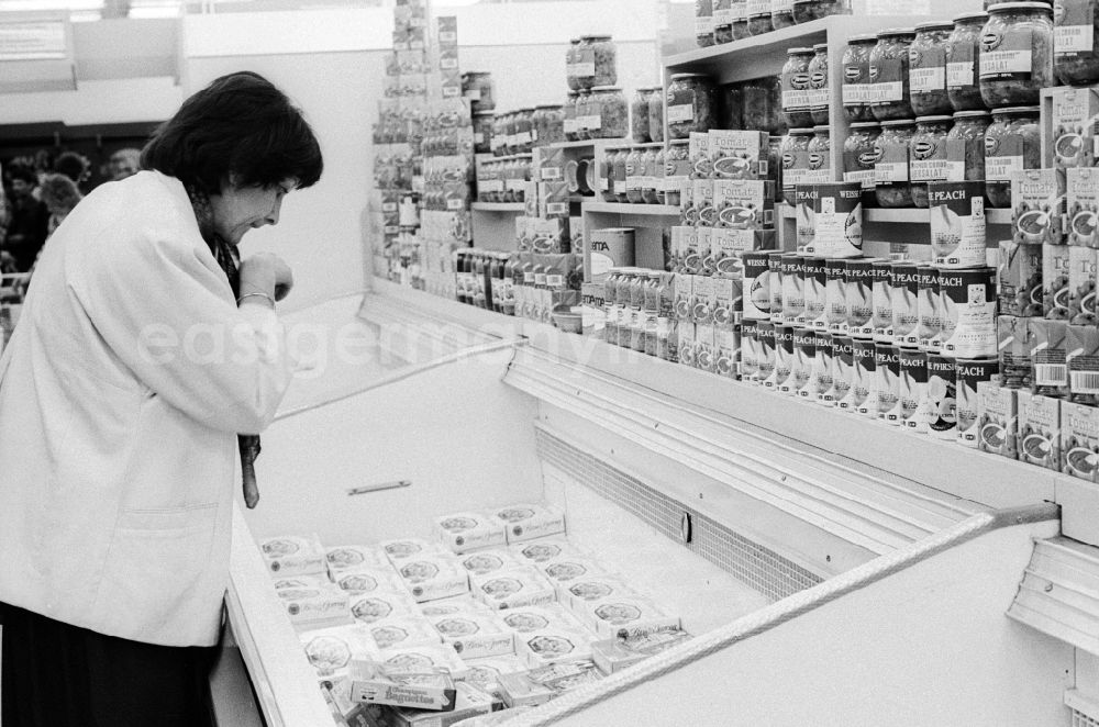 GDR photo archive: Berlin - A young woman stands in front of a freezer in a department store in Berlin, the former capital of the GDR, the German Democratic Republic. The shelves are filled partly with Ostprodukten as well with Western products