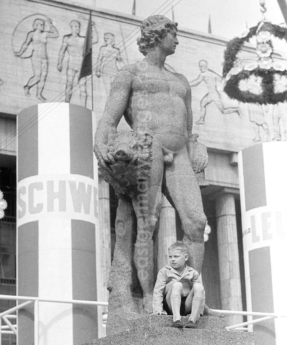 GDR photo archive: Berlin - Zu Pfingsten 1964, vom 16.-18. Mai, findet in Berlin das letzte Deutschlandtreffen der Jugend für Frieden und Völkerfreundschaft statt. Hier im Bild am Rande einer Veranstaltung auf der Karl-Marx-Allee festgehalten - ein Junge / Kind sitzt auf dem Sockel einer Steinfigur / Skulptur, die einen nackten Heroen darstellt. In seiner Hand hält die Figur ein Wildschwein-Kopf. Rechts im Bild ist ein Richtkranz zu sehen, sowie im Hintergrund ein Wandfries / Relief mit sozialistischen Motiven rund um das Thema sportliche Betätigung / Sport. Ziel der ersten Veranstaltung im Mai 1950 war, die kommunistische Jugend in Ost und West mobil zu machen und der damals in der BRD noch zugelassenen FDJ neue Anhänger zuzuführen. 1951 wurde das Verbot gegen die FDJ in der BRD ausgesprochen. Nach 1964 entsprach eine gesamtdeutsche kommunistische Politik nicht mehr den Zielen der DDR-Regierung. In den 70er und 8