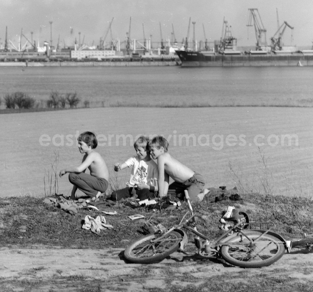 GDR image archive: Rostock - Children who have taken off their bicycles are sitting at the edge of the field in Rostock in the federal state Mecklenburg-Western Pomerania on the territory of the former GDR, German Democratic Republic. In the background the overseas port of Rostock
