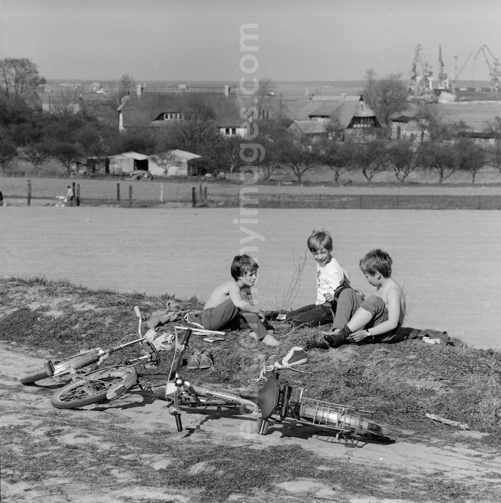 GDR photo archive: Rostock - Children who have taken off their bicycles are sitting at the edge of the field in Rostock in the federal state Mecklenburg-Western Pomerania on the territory of the former GDR, German Democratic Republic. In the background the overseas port of Rostock