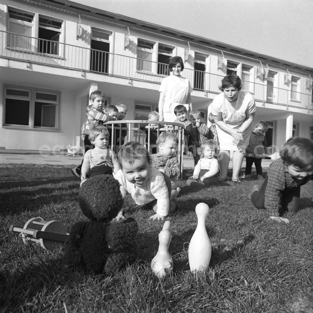 GDR picture archive: Cottbus - Kleinkinder / Kinder im Kindergarten / Kinderkrippe spielen auf der Wiese, Erzieher / Erzieherinnen betreuen die Kinder im Hintergrund
