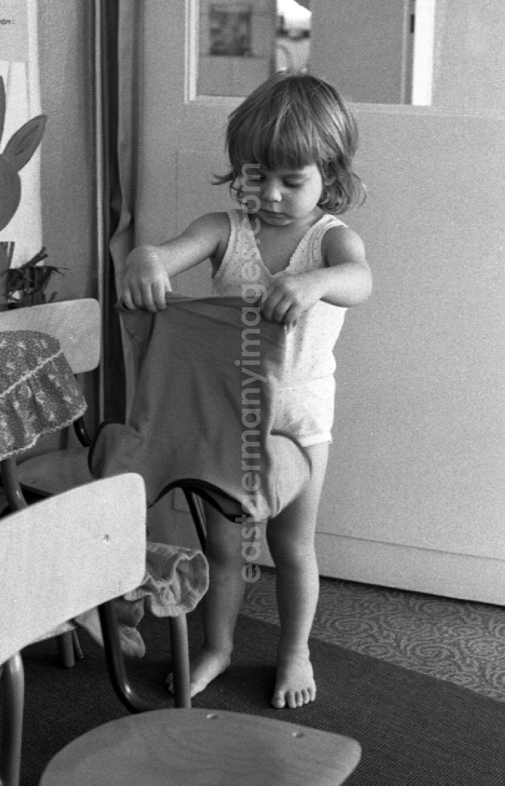 GDR picture archive: Berlin - Preparing for a nap in the kindergarten in Berlin Eastberlin on the territory of the former GDR, German Democratic Republic. Toddler tries to undress on his own and pulls on his sock or stocking. Toddler undresses and puts a T-shirt over a chair