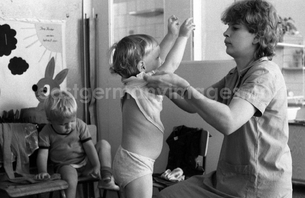 GDR image archive: Berlin - Preparing for a nap in the kindergarten in Berlin Eastberlin on the territory of the former GDR, German Democratic Republic. Nursery nurse helping a toddler undress