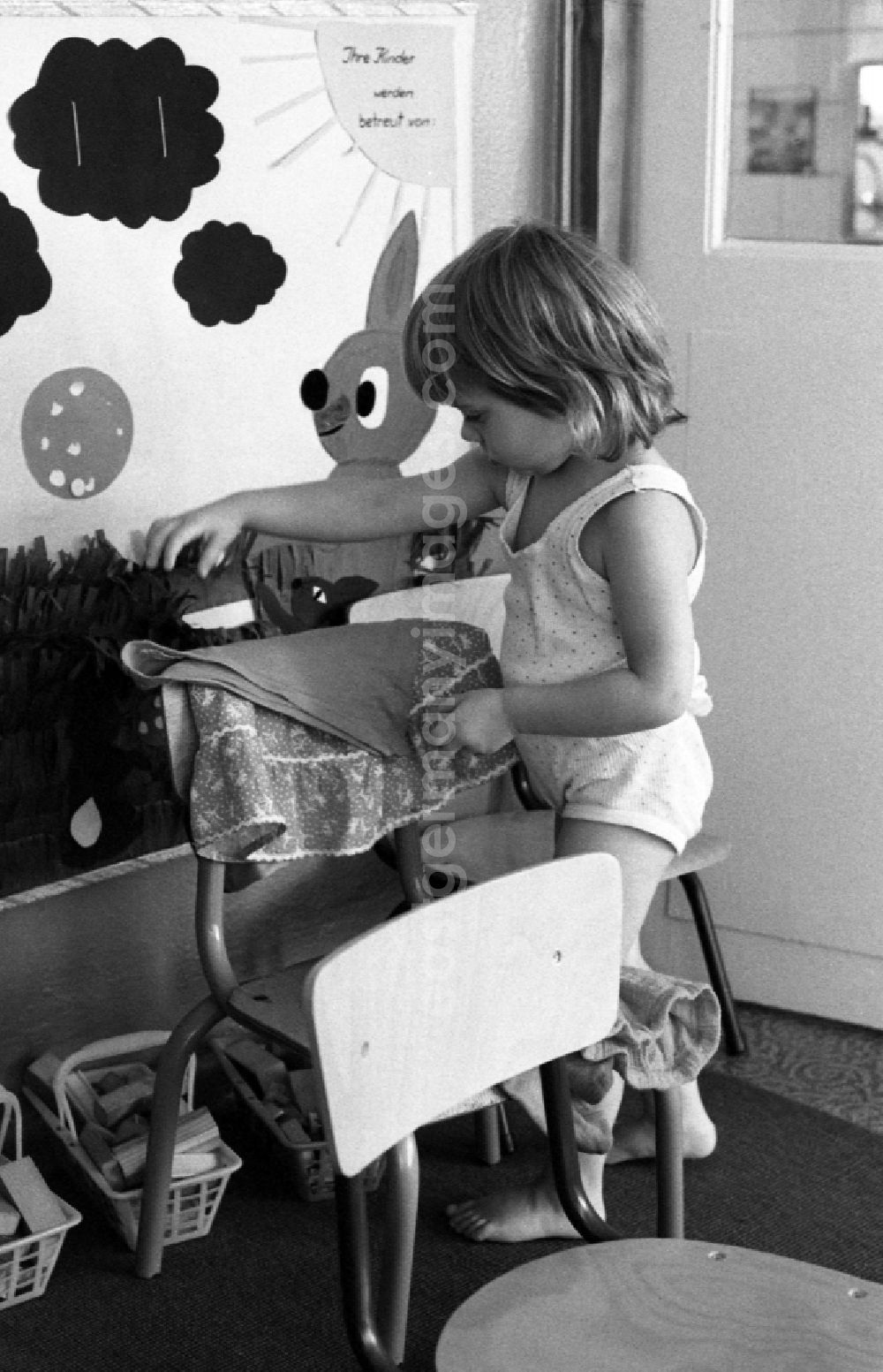 Berlin: Preparing for a nap in the kindergarten in Berlin Eastberlin on the territory of the former GDR, German Democratic Republic. Toddler tries to undress on his own and pulls on his sock or stocking. Toddler undresses and puts a T-shirt over a chair