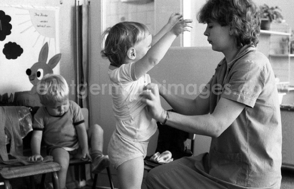 GDR photo archive: Berlin - Preparing for a nap in the kindergarten in Berlin Eastberlin on the territory of the former GDR, German Democratic Republic. Nursery nurse helping a toddler undress