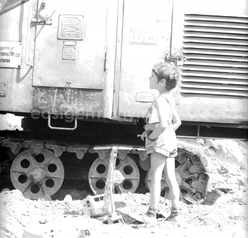 GDR photo archive: Berlin - Small boy in leather trousers plays with his wooden excavator on a building site beside a great excavator in Berlin, the former capital of the GDR, German democratic republic