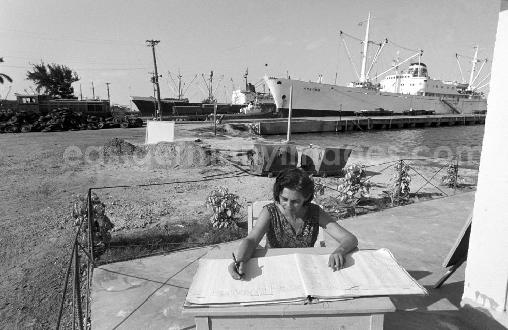 GDR picture archive: Nuevitas - Am Hafen von Nuevitas kontrolliert eine Zollangestellte den Warenverkehr. Im Hintergrund liegt das sowjetische Frachtschiff Aragwi vor Anker. In den 60er und 7