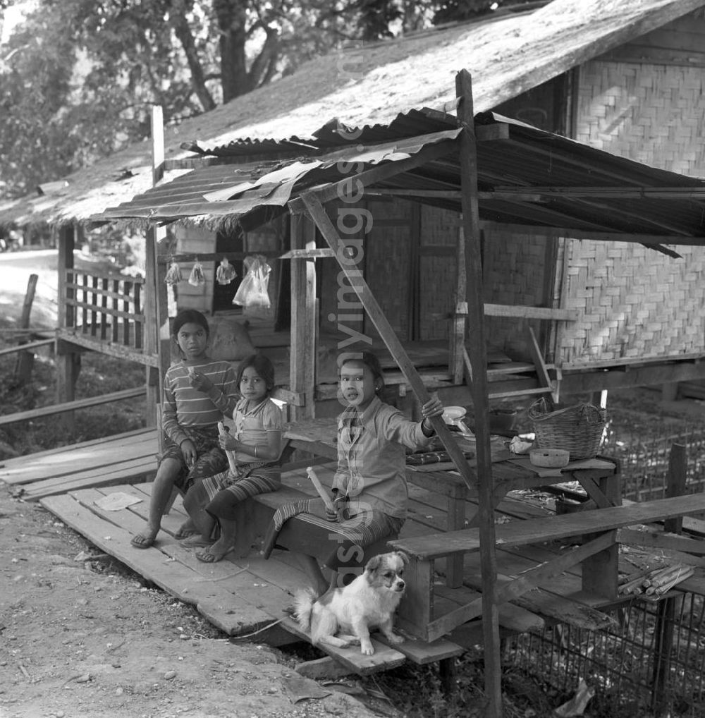 GDR image archive: Vientiane - Die Kinder einer Familie sitzen am Eßplatz vor ihrem Haus in einem Dorf nördlich von Vientiane, der Hauptstadt der Demokratischen Volksrepublik Laos.