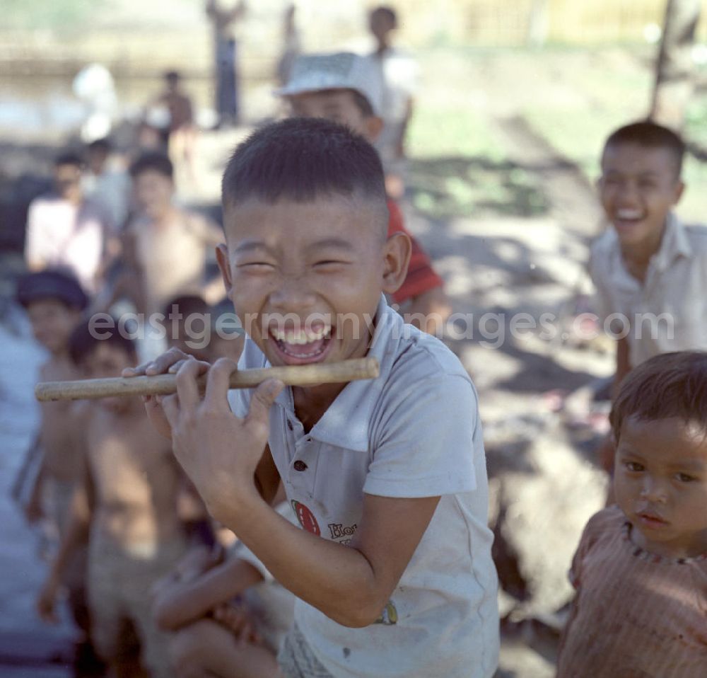 GDR picture archive: Vientiane - Lachender Junge mit Querflöte in Vientiane, der Hauptstadt der Demokratischen Volksrepublik Laos. Die Musik begleitet die anderen Schulkinder beim Bau eines Fischteichs.