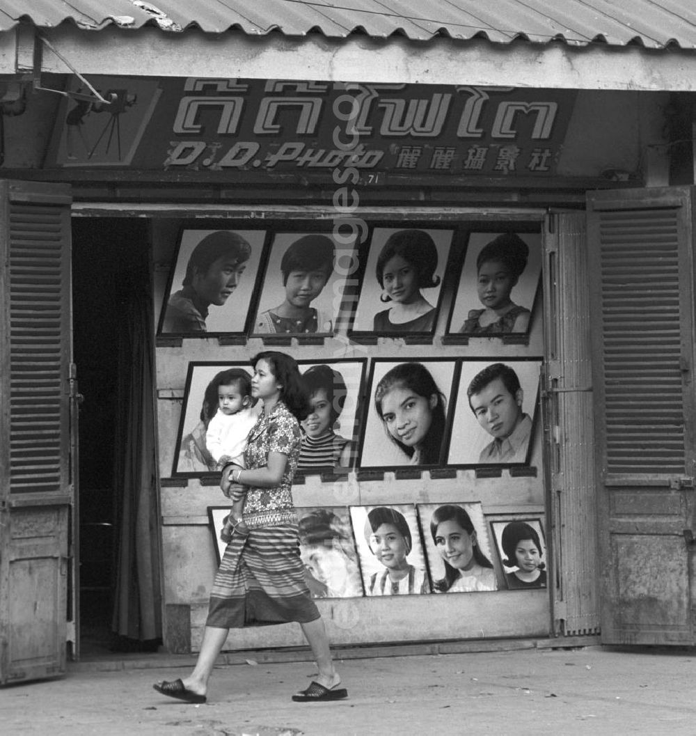 Vientiane: Eine Frau läuft mit ihrem Kind auf dem Arm an einem Fotogeschäft in Vientiane, der Hauptstadt der Demokratischen Volksrepublik Laos, vorbei, das mit großen Porträtaufnahmen im Schaufenster wirbt.