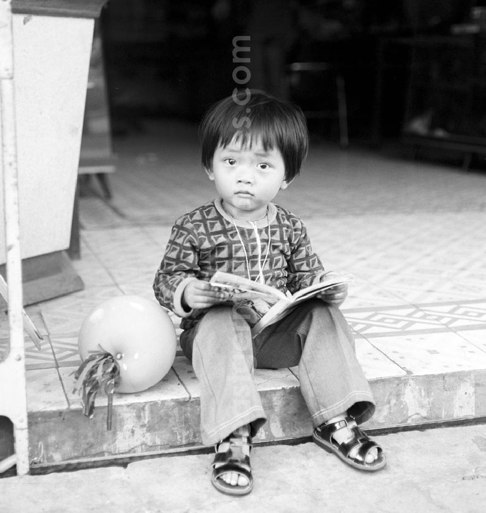 GDR photo archive: Vientiane - Ein kleiner Junge sitzt am Straßenrand auf einer Bordsteinkante und betrachtet ein Comicbuch in Vientiane, der Hauptstadt der Demokratischen Volksrepublik Laos.