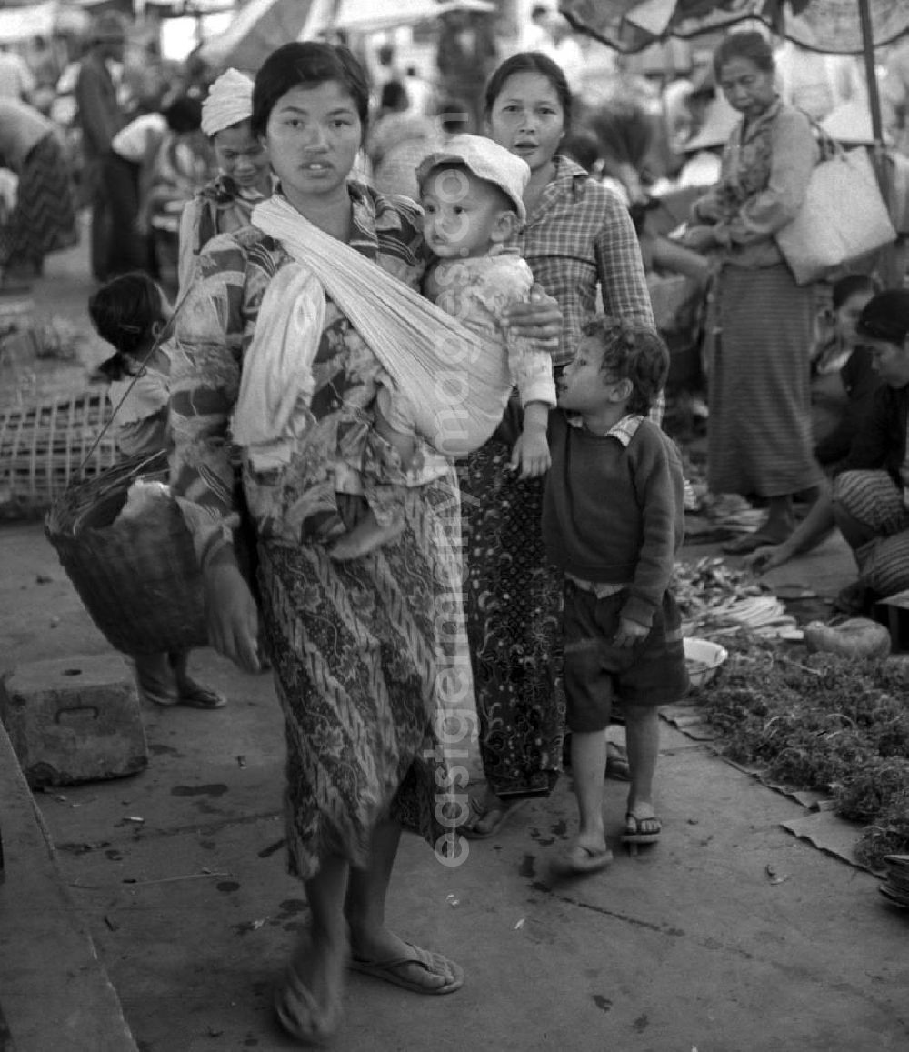 GDR picture archive: Vientiane - Eine Frau trägt ihr Baby in einem Tragetuch über den Markt in Vientiane, der Hauptstadt der Demokratischen Volksrepublik Laos.