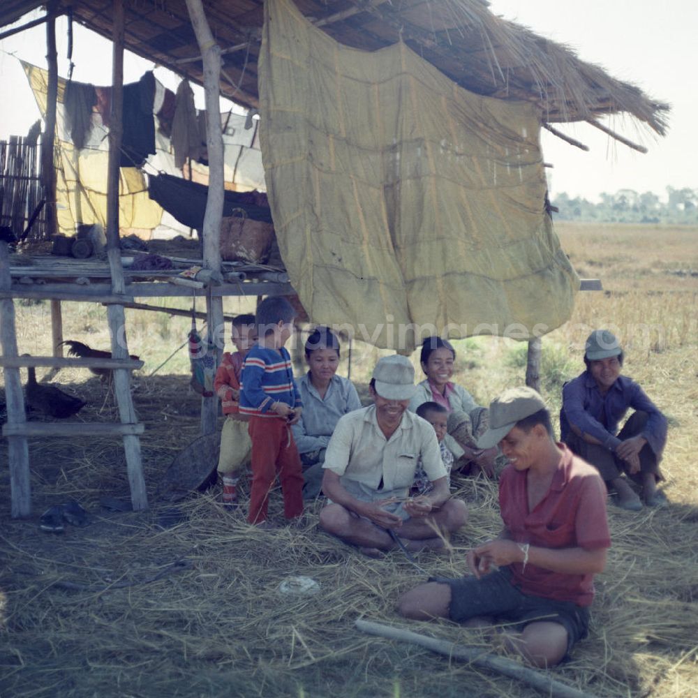 GDR photo archive: Vientiane - Männer, Frauen und Kinder sitzen während der Reisernte im Schatten einer Hütte auf einem Feld in der Demokratischen Volksrepublik Laos.