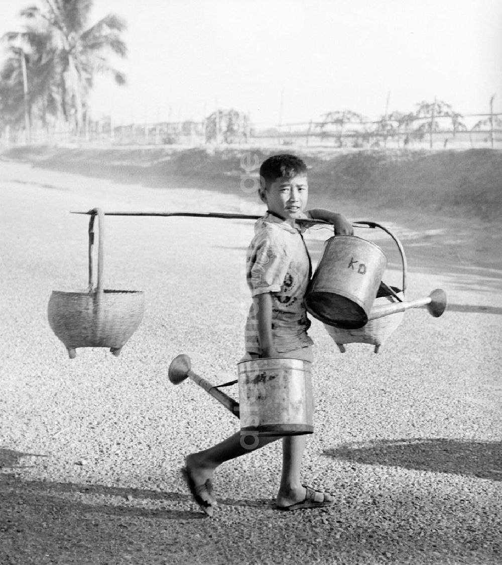 GDR image archive: Vientiane - Ein Junge trägt Gießkannen und geschulterte Körbe auf einer Straße in Vientiane, der Hauptstadt der Demokratischen Volksrepublik Laos.