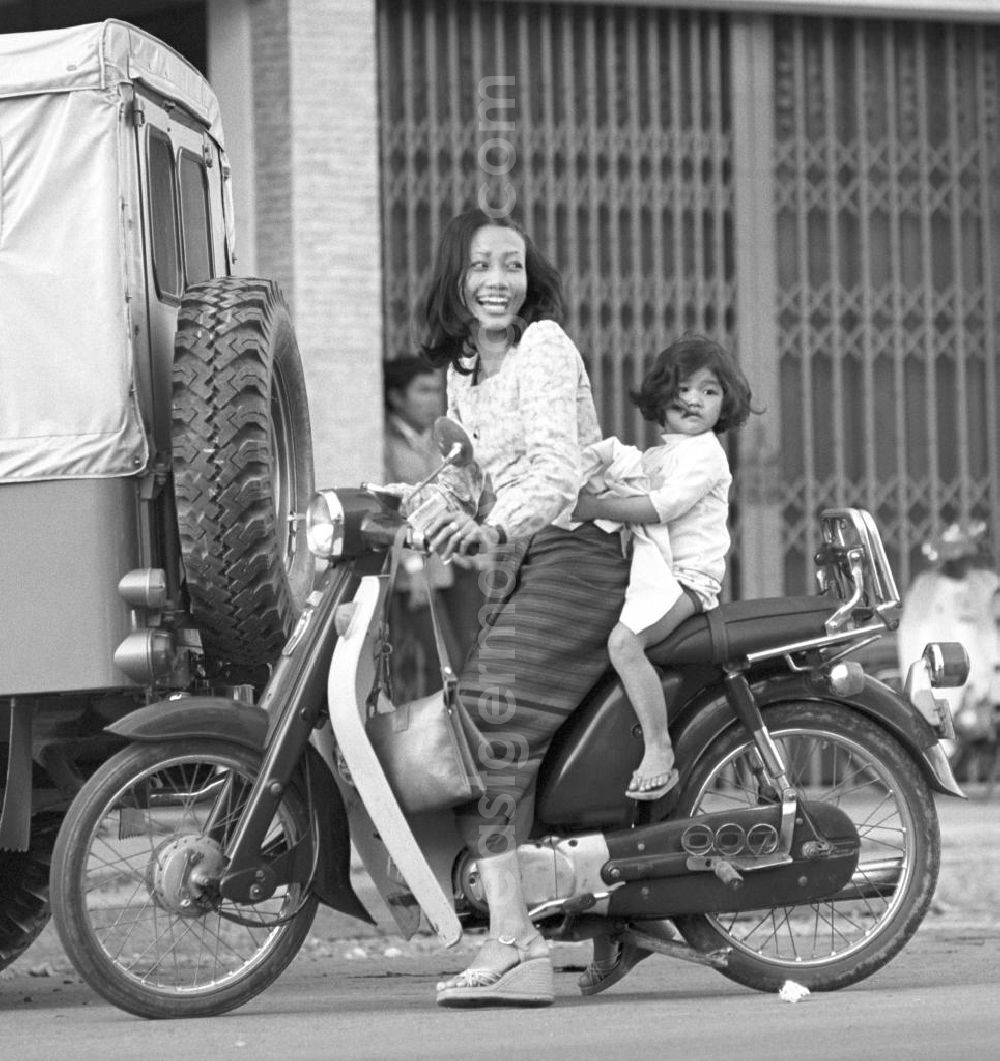 GDR photo archive: Vientiane - Eine Frau fährt mit ihrem Kind auf einem Moped auf einer Straße in Vientiane, der Hauptstadt der Demokratischen Volksrepublik Laos.
