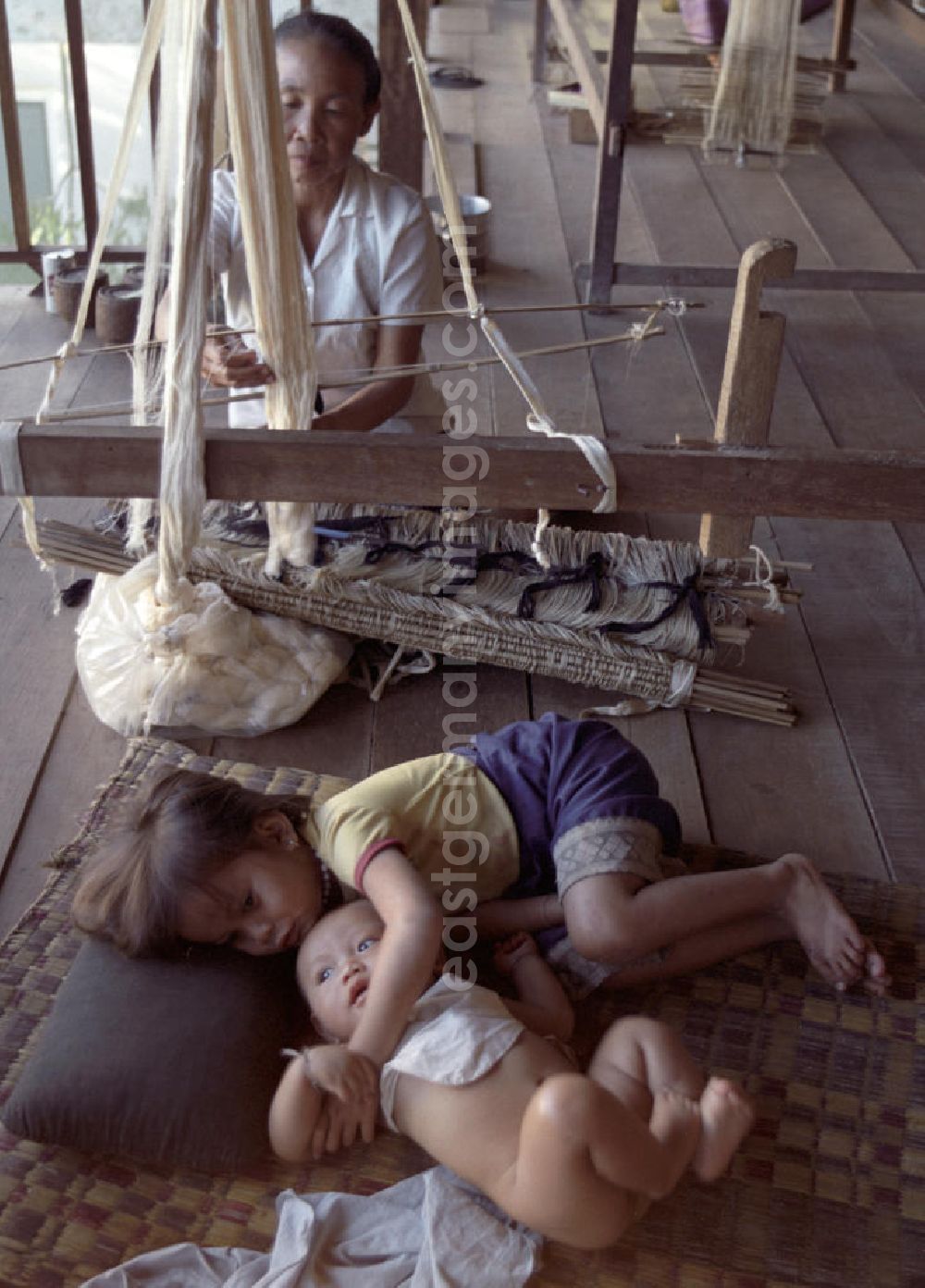 GDR image archive: Vientiane - Eine Weberin bei ihrer Arbeit, während die Kinder daneben auf einer Matte liegen in einer Weberei in Vientiane, der Hauptstadt der Demokratischen Volksrepublik Laos.