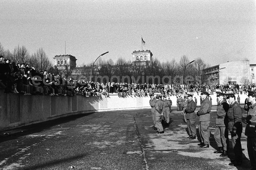 GDR picture archive: Berlin - Soldaten der DDR stehen in einer Reihe vor der Berliner-Mauer am Brandenburger Tor, auf der Mauer sitzen und stehen Menschen. Im Hintergrund der Reichstag.