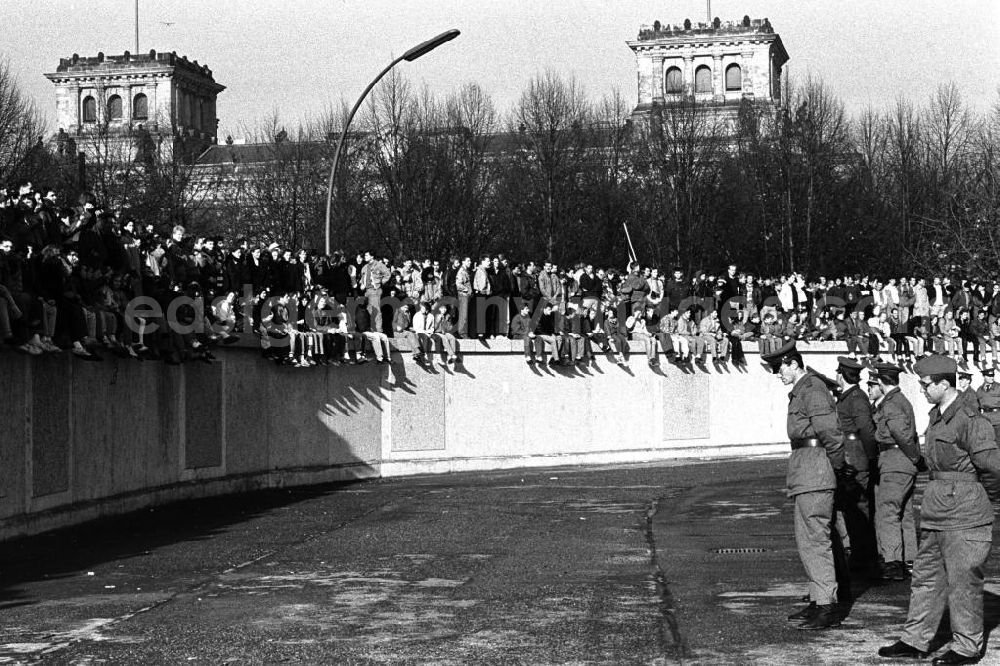 Berlin: Soldaten der DDR stehen in einer Reihe vor der Berliner-Mauer am Brandenburger Tor, auf der Mauer sitzen und stehen Menschen. Im Hintergrund der Reichstag.