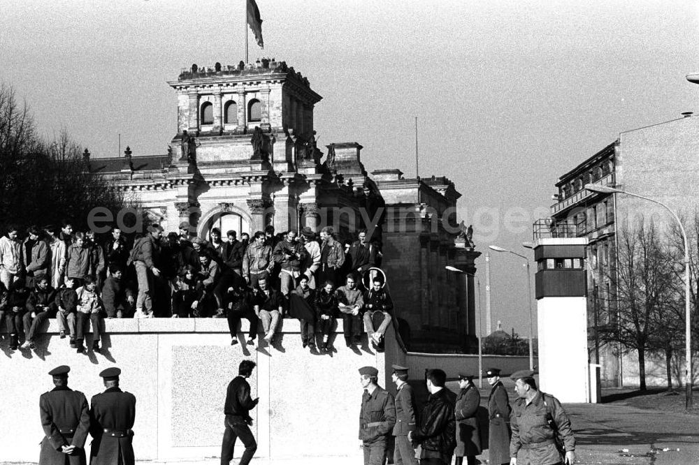 GDR photo archive: Berlin - Soldaten der DDR stehen vor der Berliner-Mauer am Brandenburger Tor, auf der Mauer sitzen und stehen Menschen. Im Hintergrund der Reichstag und ein Wachturm.