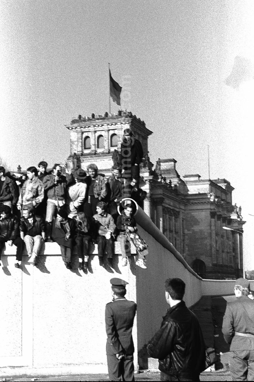 GDR picture archive: Berlin - Soldaten der DDR stehen vor der Berliner-Mauer am Brandenburger Tor, auf der Mauer sitzen und stehen Menschen. Im Hintergrund der Reichstag.