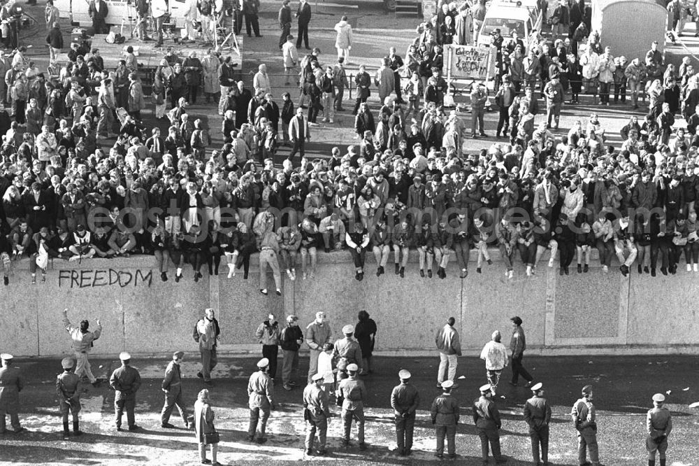 GDR picture archive: Berlin - Soldaten der DDR stehen in einer Reihe vor der Berliner-Mauer am Brandenburger Tor, auf der Mauer sitzen und stehen Menschen.