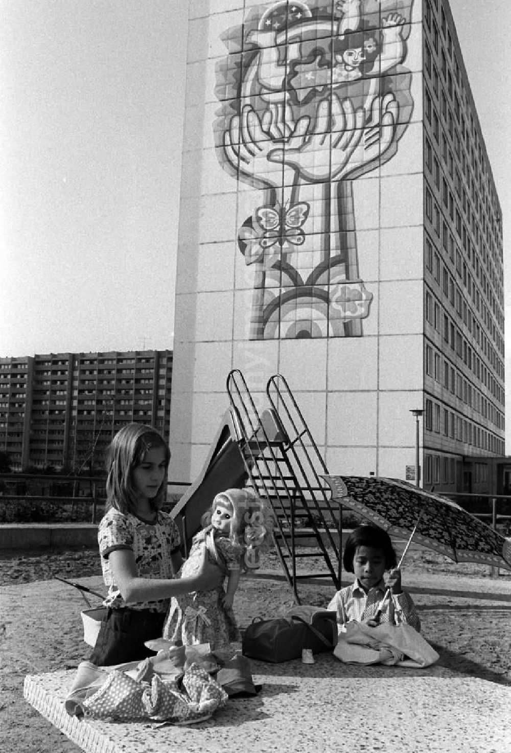 GDR picture archive: Berlin - Mädchen spielen mit Puppen auf einem Spielplatz an einem Neubaugebiet in Berlin. Ein Mädchen hält eine Schirm in der Hand. An der Fassade des elfgeschossigen Wohnhauses im Hintergrund ist ein großflächiges Bild angebracht, das schützende Hände und das Glück der Kinder auf der ganzen Welt symbolisiert.