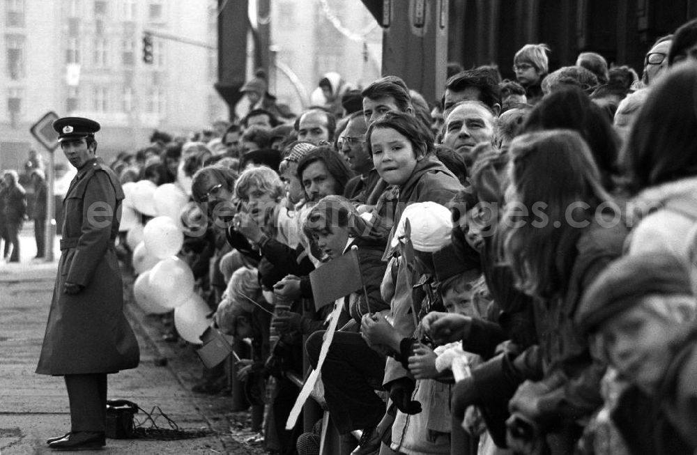 GDR image archive: Berlin - Die Bevölkerung / Menschen stehen hinter Absperrung am Straßenrand zusammen und warten auf die bevorstehende Militärparade auf der Karl-Marx-Alle anlässlich des 25-jährigen Bestehen der DDR. Kinder halten Fahnen / Winkelemente in ihren Händen. Ein Volkspolizist mit Uniform steht vor der Absperrung.