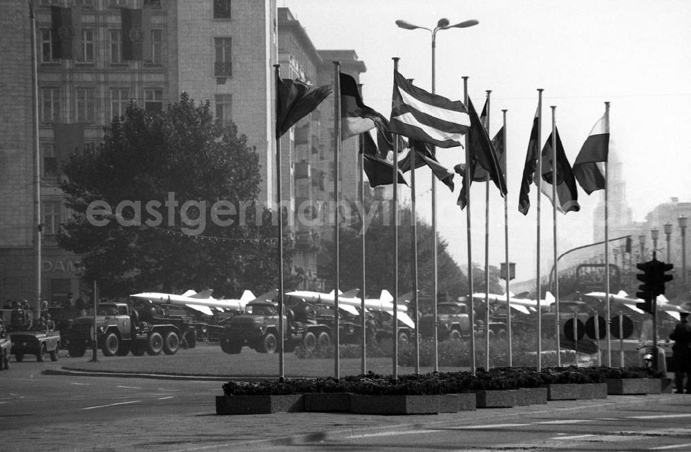 GDR photo archive: Berlin - Flaggen verschiedener solzialistischer bzw. kommunistischer Staaten, wie Kuba, Polen, DDR, Sowjetunion usw. auf dem Strausberger Platz. Im Hintergrund fahren Militärfahrzeuge mit Raketen auf der Laderampe auf der Straße anlässlich des 25-jährigen Bestehen der DDR.