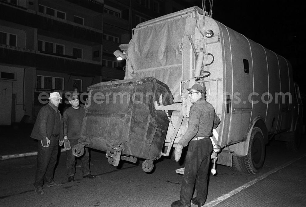 GDR picture archive: Berlin - Nachtaufnahme: Müllmänner der Müllabfuhr bei der Arbeit, Mülltonne hängt an Müllwagen / Müllfahrzeug, Müllmänner stehen daneben.