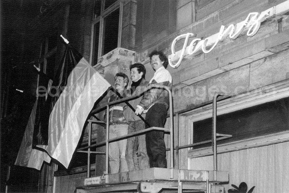 GDR picture archive: Leipzig - Demonstranten stehen zusammen auf einen Gerüst vor Flaggen der BRD. Dahinter ein Leuchtreklame mit der Aufschrift Tanz. Der Wunsch nach einem wiedervereinigten Deutschland bestimmte die Leipziger Montagsdemonstration. Die politische Eigenständigkeit der DDR wurde nur noch auf wenigen Transparenten gefordert.