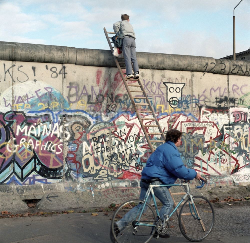 Berlin - Mitte: Neugieriger Mann klettert mit Hilfe einer Leiter an der Berliner Mauer hoch. Die Berliner Mauer war während der Teilung Deutschlands ein hermetisch abriegelndes Grenzbefestigungssystem der Deutschen Demokratischen Republik.