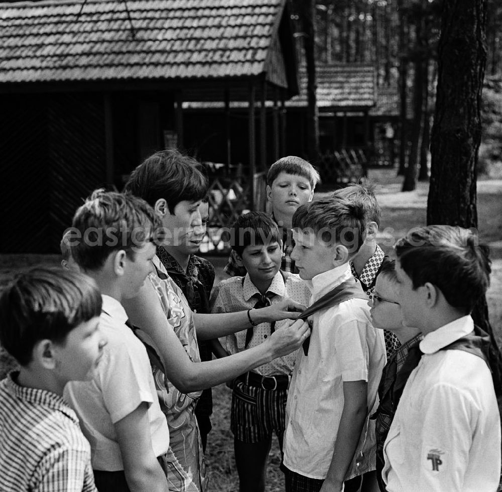 GDR picture archive: Bad Saarow - Vorbereitung auf den Fahnenappell im Ferienlager Feliks Dzierzynski in Bad Saarow. Erzieherin bindet einen Knoten mit dem Halstuch eines Pionier, Pioniere drumherum schauen zu. In der DDR gab es vor allem in den Sommerferien Betriebsferienlager, die hauptsächlich von Großbetrieben für Kinder der Beschäftigten unterhalten wurden. Außerdem gab es Pionierlager, die über die Schulen in Zusammenarbeit mit der Pionierorganisation und der FDJ organisiert wurden.