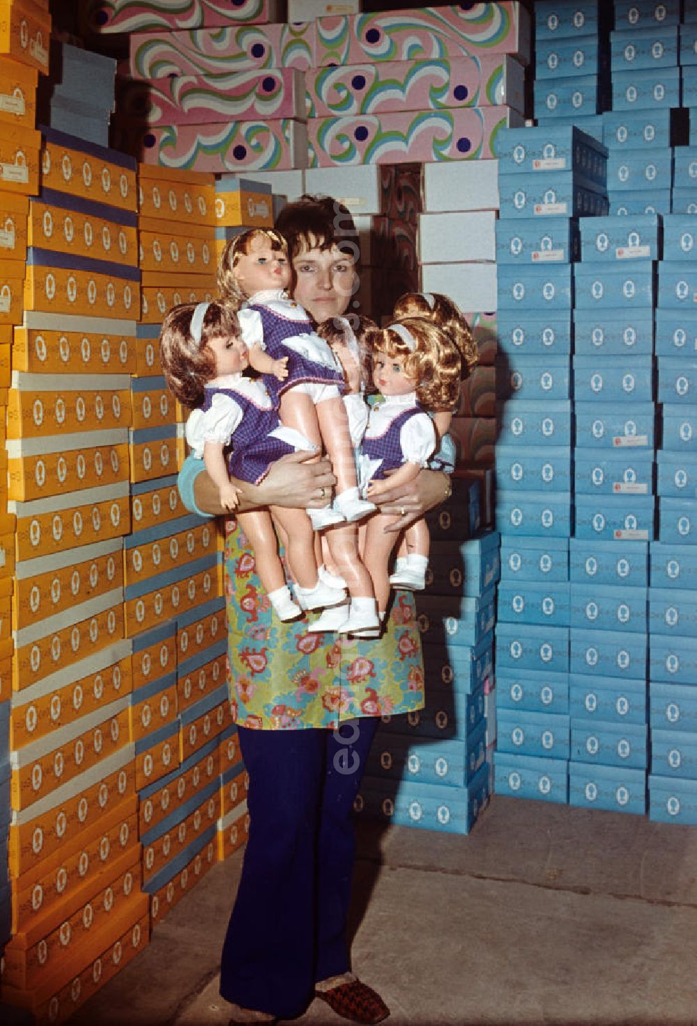 GDR image archive: Sonneberg - Eine Mitarbeiterin des VEB Kombinat Spielwaren Sonneberg sonni präsentiert fertige Puppen auf ihrem Arm, im Hintergrund stapeln sich die Verkaufskartons. Die Sonneberger Puppen waren nicht nur in der DDR beliebt, sondern trugen den Namen der Stadt in alle Welt.