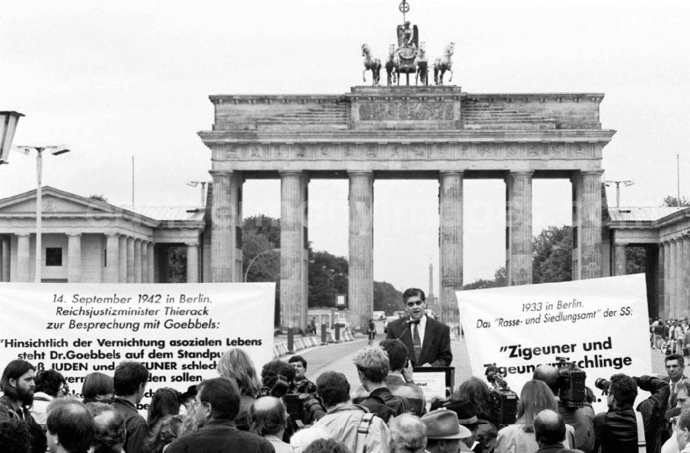 GDR picture archive: Berlin-Mitte - Sintis und Roma demonstrieren auf dem Pariser Platz vor dem Brandenburger Tor. Mann steht an Rednerput, davor Pressevertreter (Rückansicht). Demonstranten halten Plakate.